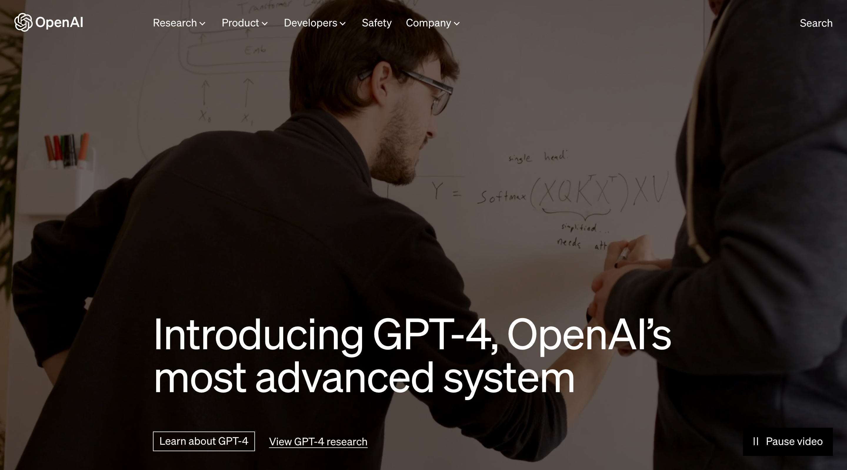 Screenshot of the OpenAI website hero. The background is an auto-playing video showing people working on different things in an office. There’s a dark overlay on top of the video, and the website text and OpenAI logo are white. In the corner is a button to pause the video. The headline is at the bottom of the hero with a primary and secondary button below it. The navbar is at the top and has a transparent background. It contains the OpenAI logo, dropdown lists and links, and a search button.