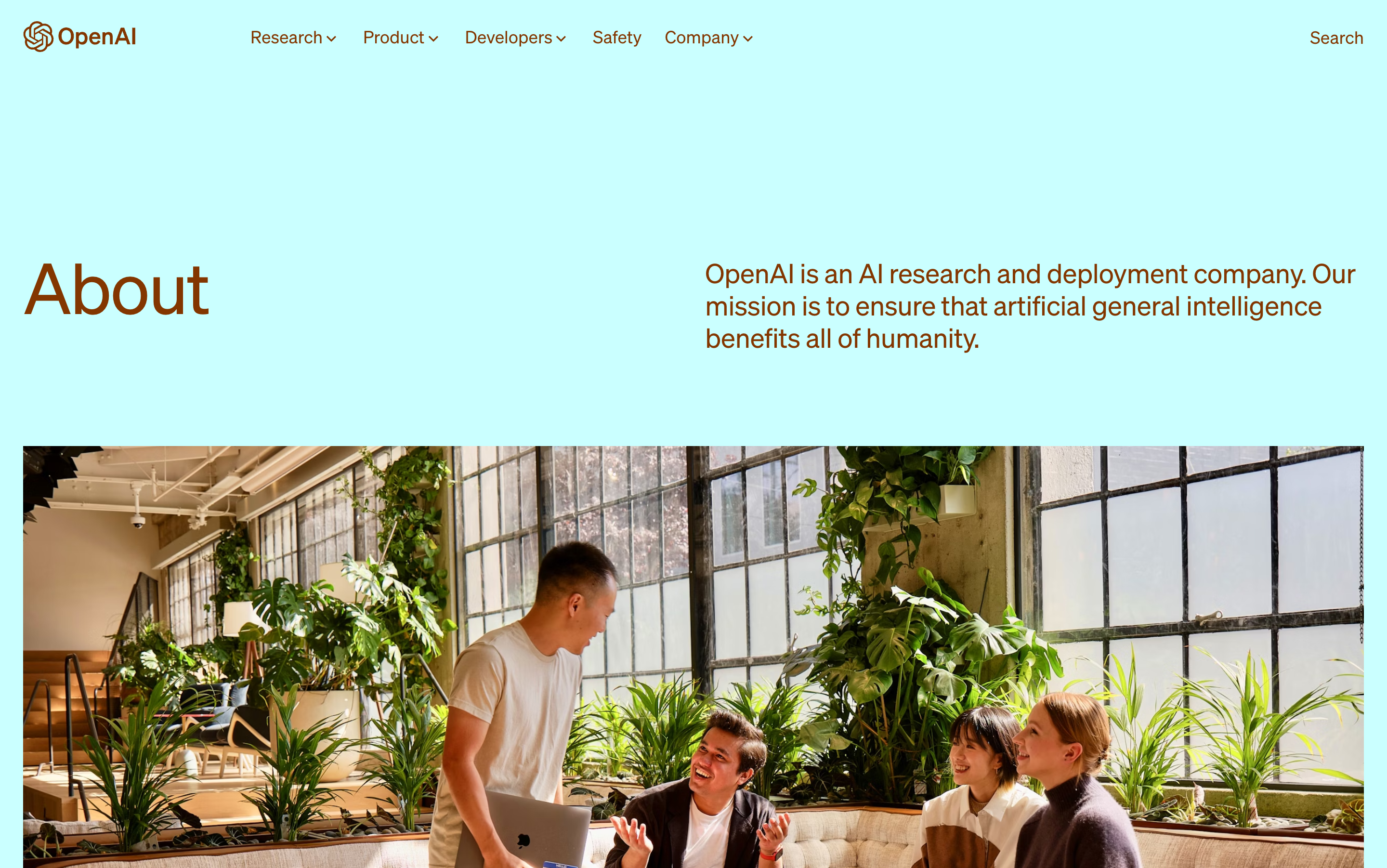 Screenshot of the hero section of the OpenAI website about page. The headline is to the left, and the subheadline is to the right. Below is a full-width image showing four happy people in an office that looks like an old factory floor with much daylight and many green plants. The page's background color is light cyan, and the text color is dark orange.